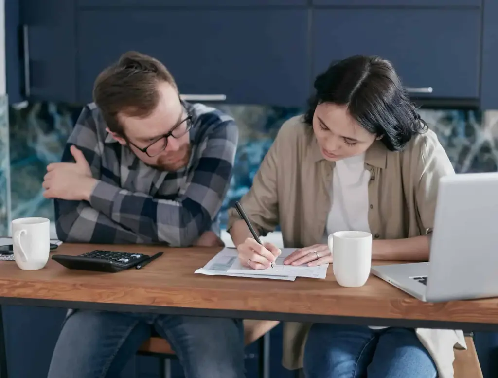 A woman assists a man in filling out paperwork while they sit together at a table with laptops.