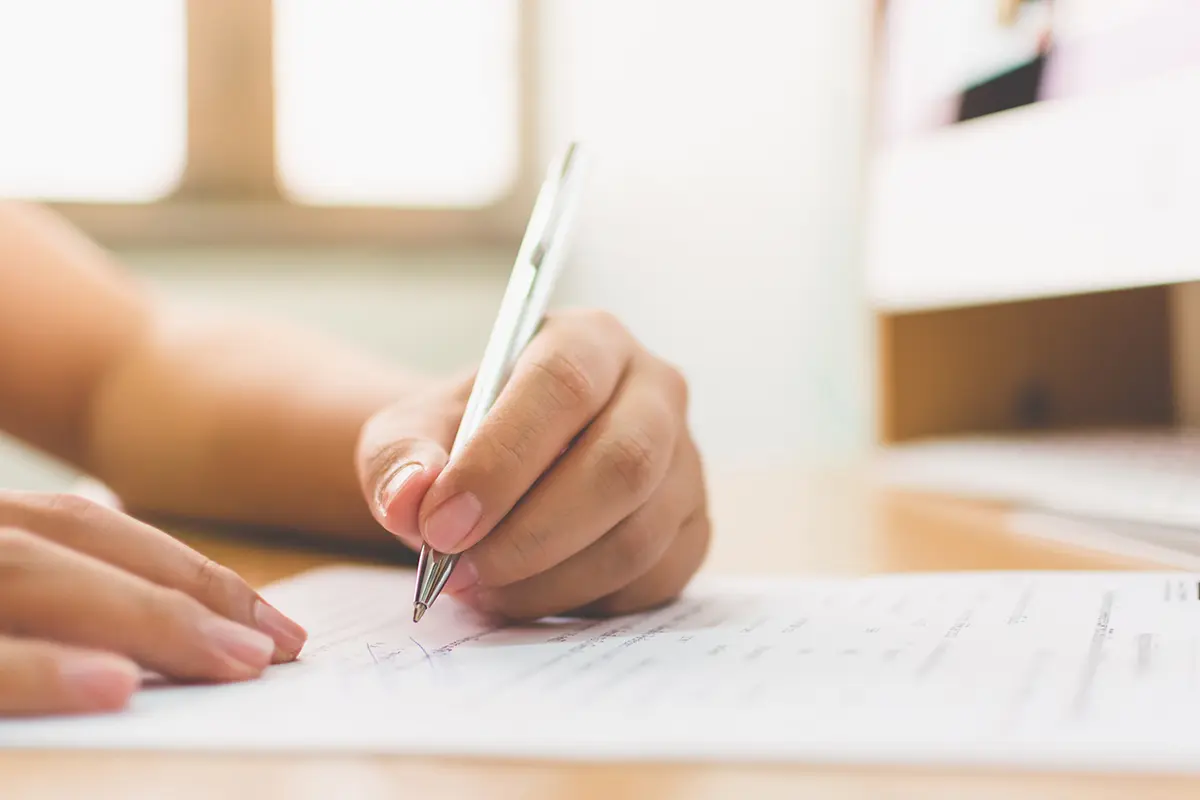 A close-up view of a person's hand holding a silver pen, signing a document on a wooden desk.