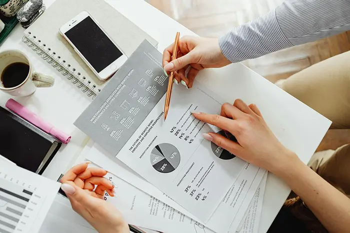 A person pointing with a pen at printed charts and business statistics on a desk with paperwork.