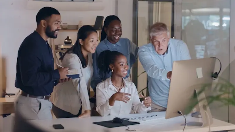 Diverse team of coworkers gathered happily around a computer screen in a modern office, collaborating on a project.