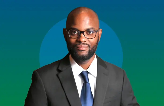 Portrait of Noel Neil wearing a dark suit, tie, and glasses against a blue and green background