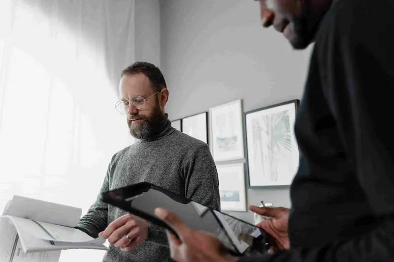 Two colleagues collaborate indoors, with one holding a printed document and the other using a digital tablet.