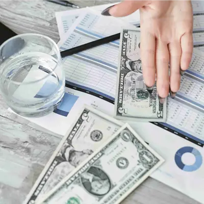 A desk with US dollar bills, financial charts, a glass of water, and a person's hand