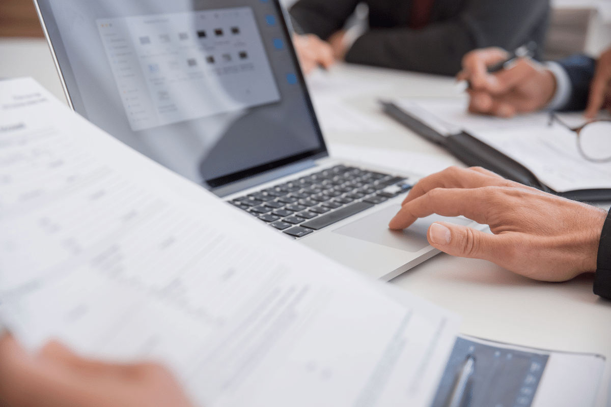 A person pointing with a pen at printed charts and business statistics on a desk with paperwork.