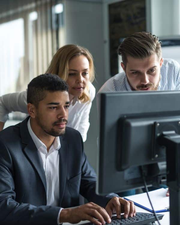 Three diverse coworkers smiling together while collaborating in front of a computer monitor and a digital tablet in an office