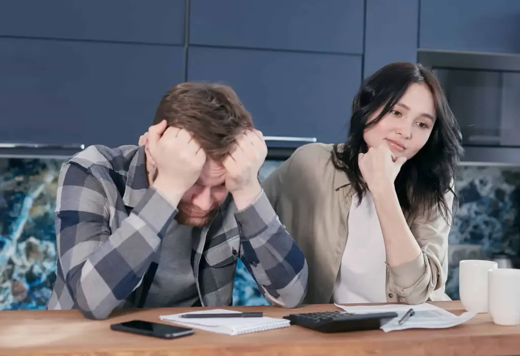 A stressed man holding his head while a concerned woman looks on, seated at a table with financial documents