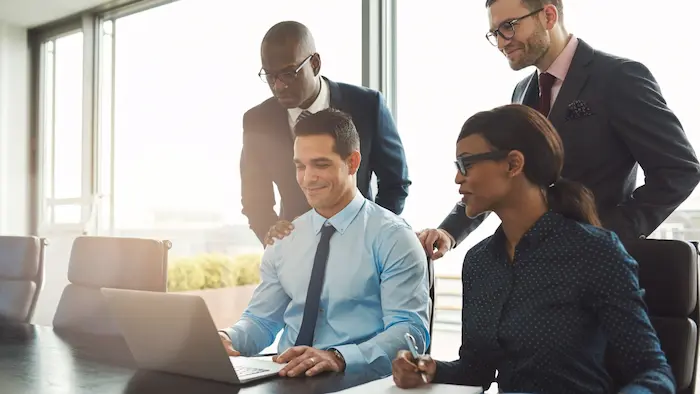 Four business professionals in suits gathering around a laptop together in a bright office conference room.